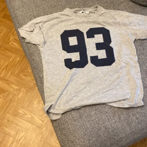 Grå t-shirt med tryck från 157 - Säljer en ljusgrå t-shirt från 157 med stort svart tryck av siffran 93 på framsidan. T-shirten har rund hals och korta ärmar, perfekt för en avslappnad stil.