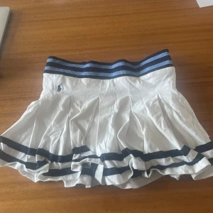 Vit plisserad kjol från Polo Ralph Lauren - Säljer en vit plisserad kjol från Polo Ralph Lauren med marinblå ränder i midjan och längs fållen. Kjolen har broderad logga framtill och bred resår i midjan för en sportig look.