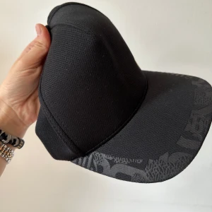 Svart BOSS keps (Hugo Boss) - Svart keps med modern design och snygga mönsterdetaljer på skärmen som ger den en unik och sofistikerad look. Den guldfärgade Hugo Boss-loggan ger en exklusiv touch. Den är i mycket gott skick, inga skador eller fläckar. Säljer den eftersom det var en present från min man, men den var inte i min smak. Perfekt för dig som uppskattar kvalitet och vill ha en stilfull accessoar