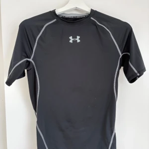 Svart compression shirt från Under Armour - Svart kortärmad compression shirt från Under Armour med gråa sömmar och logga på bröstet. Tight passform som sitter nära kroppen, perfekt för intensiva träningspass.