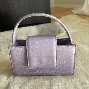 Ljuslila handväska med krokodilpräglat mönster - Säljer en ljuslila handväska i syntetmaterial med krokodilpräglat mönster. Väskan har ett kort handtag och en rektangulär form med lock framtill. Perfekt accessoar för att lyfta din outfit!