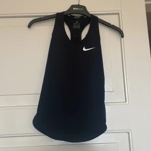 Svart träningstopp från Nike - Säljer en svart ärmlös träningstopp från Nike med Dri-Fit material och klassisk vit logga på bröstet. Perfekt för sport och träning, med racerback-design för extra rörelsefrihet.