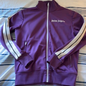 Lila track jacket från Palm Angels - Medium - Säljer en lila track jacket från Palm Angels med vita ränder längs ärmarna och dragkedja framtill. Jackan har hög krage, fickor med dragkedja och logga på bröstet. Perfekt för en sportig och trendig look. Storlek Medium