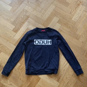 Hugo boss kofta - Säljer en mörk blå sweatshirt från Hugo med vit logga tryckt på bröstet. Tröjan har rund halsringning och långa ärmar. Perfekt för en stilren och enkel look.