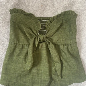 Grön axelbandslös topp med knyt från SHEIN - Snygg grön axelbandslös topp från SHEIN med volangkant upptill och knyt framtill. Toppen har smockad rygg för extra komfort och är tillverkad i 100% polyester. Perfekt för varma dagar och en trendig look.