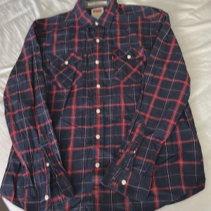 Rutig skjorta från Levi's i blått och rött - Säljer en klassisk långärmad skjorta från Levi's i blått med rött och vitt rutmönster. Skjortan har knappar framtill och två bröstfickor med lock. Perfekt för en avslappnad och stilren look.