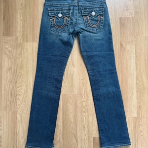 True Religion jeans med broderade fickor - Säljer ett par klassiska blå jeans från True Religion med snygga kontrastsömmar och broderade bakfickor. Byxorna har bootcut och silverfärgade knappar. Perfekta för dig som gillar detaljer och ikonisk denimstil. Skicket är 10/10 och är som nya.