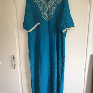 Turkos koftan med broderad detalj - Säljer en turkos långklänning med vackert vitt broderi vid halsen och mönstrad struktur i tyget. Klänningen har korta ärmar med dekorativ kant. Perfekt för dig som vill sticka ut med färg och stil.