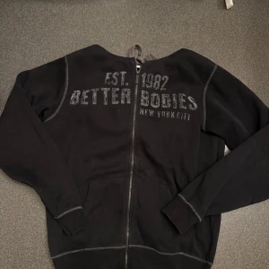 Svart hoodie från Better Bodies - Svart hoodie med dragkedja från Better Bodies. Tryck på bröstet med texten 'EST. 1982 BETTER BODIES NEW YORK CITY'. Huva och två fickor framtill. Perfekt för träning eller vardag.              Bra skick knappt använd.