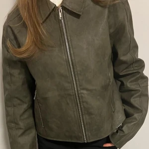  Jacka - Snygg grön jacka i skinnimitation med silvriga dragkedjor framtill och vid fickorna samt i ärmsluten. Jackan har klassisk krage och raka, långa ärmar. Passformen är normal och jackan är enkel att matcha till olika outfits. Helt oanvänd!