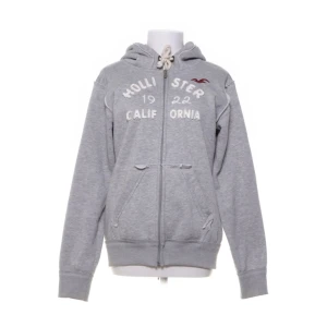 Grå hoodie från Hollister L - Köpt på Sellpy men va alldeles för liten. Står XXL på lappen men är mycket närmare en M-L i storleken. Mycket bra kvalité ändå! Grå hoodie från Hollister med dragkedja och vit broderad text framtill. Tröjan har huva med dragsko och två fickor på magen. Tillverkad i 100% bomull för en mjuk och bekväm känsla. 
