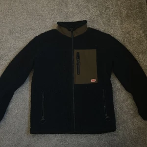 Svart fleecejacka från Armor Lux - Mysig svart fleecejacka från Armor Lux med kontrasterande olivgröna detaljer på bröstficka, axlar och krage. Jackan har dragkedja framtill, två sidofickor med dragkedja. Är använd några gånger men skicket på jackan är i princip nytt, kan inte hitta några defekter. Köparen står för frakt. Hör av dig om du har några frågor!