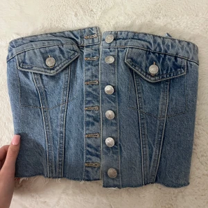 Jeans korsettopp med knappar - Säljer en snygg jeans korsettopp med silverfärgade knappar framtill och två bröstfickor med lock. Toppen är croppad med rå kant nertill och har en tight passform. Perfekt för att skapa en cool och trendig look.
