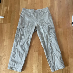 Beiga cargobyxor från W.C. Gordon - Säljer ett par beiga cargobyxor från W.C. Gordon med flera praktiska fickor på sidorna och bak. Byxorna har normal passform och är tillverkade i bomull, perfekta för en avslappnad stil.