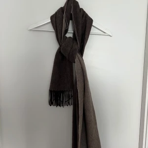 Brun halsduk från These Glory Days - Säljer en stilren brun halsduk från These Glory Days. Halsduken har klassiska fransar i ändarna och är i ett mjukt material som passar perfekt till kyliga dagar. Enkel att matcha med olika outfits.