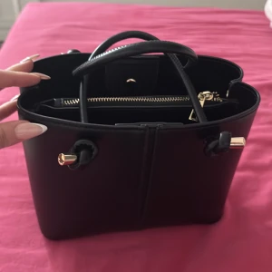 Svart handväska med gulddetaljer frän zara - Säljer en stilren svart handväska med guldfärgade detaljer och flera fack, inklusive ett med dragkedja. Väskan har två handtag och en avlång, elegant form. Perfekt för dig som vill ha en snygg accessoar till vardagslooken.