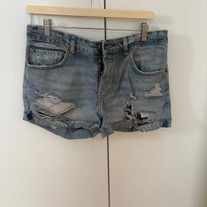 Blå lågmidjade jeansshorts med slitningar - Säljer ett par blå jeansshorts från Levi's med rejält slitna detaljer och fransig nederkant. Klassisk femficksmodell med knappgylf och hög midja. Perfekta för en avslappnad och trendig sommarstil.