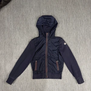 Mörkblå vindjacka med huva från Moncler - Säljer en mörkblå vindjacka från Moncler med huva och dragkedja framtill. Jackan har två fickor med dragkedja och Moncler-logga på ärmen. Snygga röda och vita detaljer längs dragkedjorna. Perfekt för säsongens alla äventyr.
