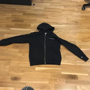 Svart hoodie med dragkedja från Calvin Klein Jeans - Svart hoodie från Calvin Klein Jeans med dragkedja framtill och huva. Diskret logga på bröstet och CK-detalj på ärmen. Perfekt för en avslappnad stil. (Junior storlek 164)
