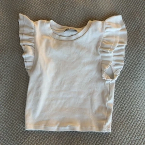Vit ribbad linnetopp med volangärmar från Zara - Supersöt vit ribbad topp från Zara med volangdetaljer på axlarna. Ärmlös modell med rund halsringning och croppad passform. Perfekt till sommaren och enkel att matcha med olika outfits.