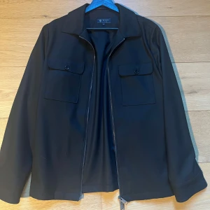 Svart jacka med dragkedja från Riley - Säljer en svart jacka från Riley med klassisk krage och två bröstfickor med knappar. Jackan har dragkedja framtill och raka ärmar. Perfekt för en stilren look. Storlek 46. 