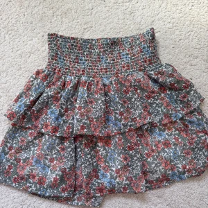 Blommig volangkjol från SRM HLT - Supersöt blommig kjol från SNR HLT med volanger och smock i midjan. Kjolen har ett färgglatt mönster i rött, blått och vitt på grå botten. Perfekt för dig som gillar en lekfull och trendig stil.