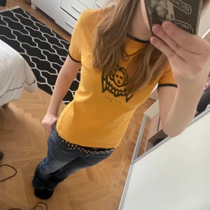 Gul t-shirt med svart tryck  - Säljer en gul t-shirt från Camera med svart grafiskt tryck framtill. T-shirten har svarta detaljer vid ärmslut och halsringning. Modellen är figurnära och har korta ärmar. Perfekt för dig som gillar unika prints! Står ingen storlek men skulle gissa på S.