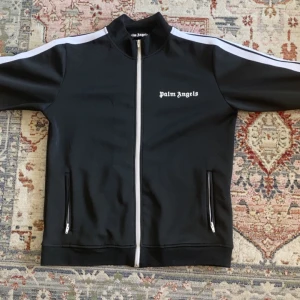 Svart track jacket från Palm Angels - Svart track jacket från Palm Angels med vit logga på bröstet och vita ränder längs ärmarna. Jackan har dragkedja framtill och två fickor med dragkedja. Hög krage och sportig look.