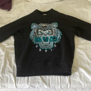 Tjocktröja från kenzo - Säljer en svart sweatshirt från Kenzo med ikonisk tigerbrodyr och texten 'KENZO PARIS' framtill. Tröjan har rund halsringning och långa ärmar med ribbade muddar. Perfekt statement-plagg för dig som gillar bra stil.