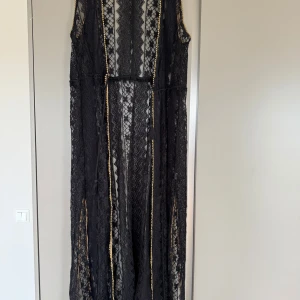 Svart lång kaftan i spets från Ibiza Flower - Säljer en svart kaftan i spets från Ibiza Flower. Klänningen är ärmlös och har dekorativa guldfärgade detaljer längs kanterna. Perfekt för dig som vill sticka ut med en elegant och bohemisk look. Perfekt till stranden. Den har ingen storlek men skulle säga att den passar 36/38