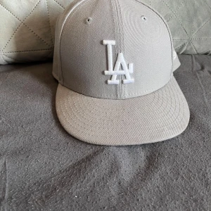 Beige LA-keps från New Era - Snygg beige keps från New Era med LA-logga broderad framtill i vitt. Klassisk modell med böjd skärm och ventilation på ovansidan. Perfekt accessoar för en stilren och sportig look.