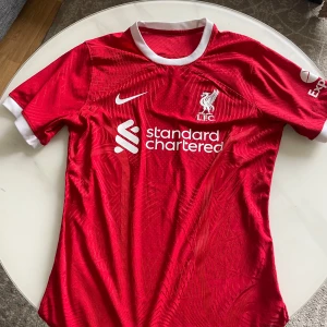 Liverpool FC hemmatröja 2023/2024 - Säljer en röd Liverpool FC hemmatröja från Nike med vita detaljer och klubbmärke på bröstet. Tröjan har korta ärmar och sponsorlogga framtill. Perfekt för dig som älskar fotboll och vill visa ditt stöd för LFC!