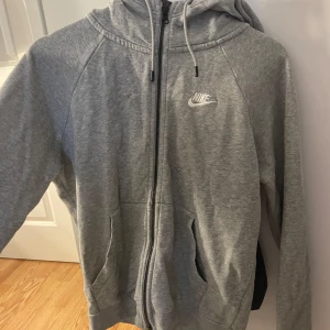 Grå zip-hoodie från Nike - Grå hoodie från Nike med dragkedja framtill och vit logga på bröstet. Jackan har huva med dragsko och två fickor framtill. Perfekt för en sportig och avslappnad stil.
