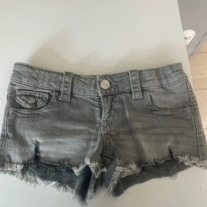 Grå jeansshorts - Säljer ett par grå jeansshorts med fransig kant och snygga detaljer som fickor med lock och silverfärgade knappar. Shortsen har bälteshällor och klassisk femficksdesign. Perfekta för en avslappnad stil.