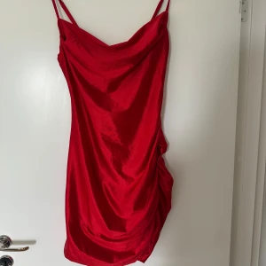Röd satin klänning från Bik Bok - Säljer en röd kortklänning i satin från Bik Bok med smala axelband och draperad effekt på sidan. Klänningen har en snygg cowlneck-ringning och är perfekt för festliga tillfällen. Sitter tight och har en glansig finish.
