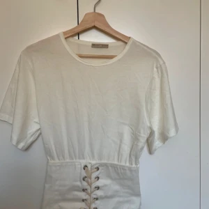 Vit topp med snörning i midjan - Säljer en vit topp med korta ärmar och rund halsringning. Toppen har en snygg knytning där framme och sitter tajt runt midjan som en corset! Tryck inte på köp nu