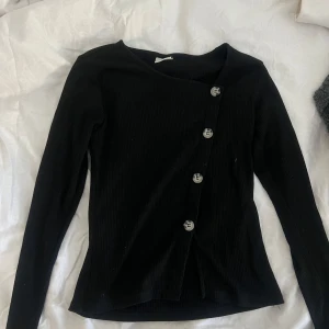 Svart ribbad topp med sned knäppning - Säljer en svart långärmad ribbad topp med sned knäppning framtill och fyra stora knappar. Toppen har en asymmetrisk halsringning och sitter figurnära. Perfekt som fest topp eller till vardags