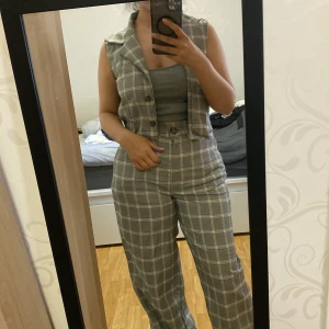 Grå rutig väst - Snygg grå väst med rutigt mönster och klassisk krage. Västen har knappar framtill och är ärmlös, perfekt att styla över en topp eller skjorta för en trendig look.