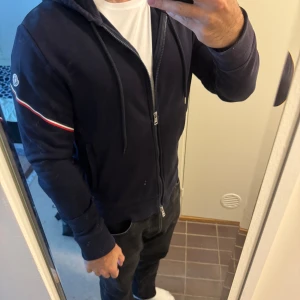 Mörkblå hoodiejacka från Moncler - Säljer en mörkblå hoodiejacka från Moncler med dragkedja och huva. Jackan har en vit och röd rand längs ärmen samt Moncler-logga på axeln. Perfekt för dig som vill ha en stilren och sportig look.