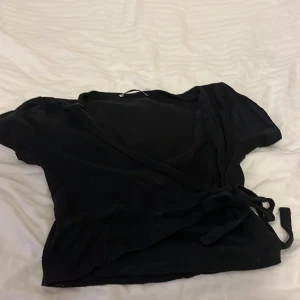 Svart omlottopp från FB Sister - Säljer en svart omlottopp från FB Sister i storlek XXS. Toppen har korta ärmar och knyt i sidan för en snygg passform. Perfekt till många olika outfits och enkel att matcha med andra plagg.