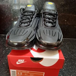 Nike Tuned Air svarta sneakers - Säljer ett par svarta Nike Tuned Air sneakers med sportig design och tydliga detaljer på ovandelen. Skorna har snörning och en robust, platt sula med synliga luftkuddar. Perfekta för dig som gillar en modern och stilren look.