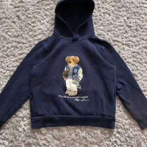 Mörkblå hoodie med björn från Polo Ralph Lauren - Säljer en mörkblå hoodie från Polo Ralph Lauren med en ikonisk björn på framsidan. Tröjan har huva och lång ärm, samt tryck med björn i sportig outfit. Perfekt för dig som gillar klassisk och sportig stil.
