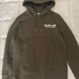 Helmut Lang hoodie - Säljer en olivgrön hoodie från Helmut Lang med vit logga tryckt på bröstet. Tröjan har huva med dragsko och en stor magficka framtill. Perfekt för dig som gillar stilrena och bekväma plagg. Made in Portugal 🇵🇹. Nypris:4500kr