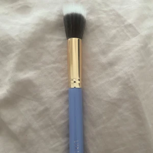 LUXIE sminkborste med blått handtag - Säljer en sminkborste från LUXIE med blått handtag och guldfärgat skaft. Borsten har täta, tvåfärgade borststrån i svart och vitt, perfekt för applicering av foundation eller highlighter.