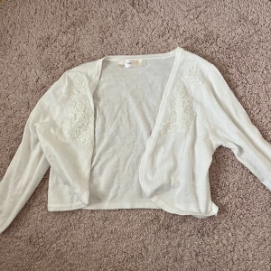 Vit bolerotopp - Säljer en vit bolerotopp från bpc selection i storlek M. Toppen har lång ärm och fina broderade blommor framtill. Perfekt att slänga över en klänning eller topp för en extra touch. Använd några enstaka gånger!