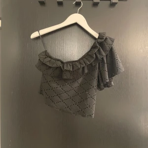 Svart offshoulder topp från Zara - Snygg svart offshoulder topp från Zara med volangdetaljer och ett hålmönster. Perfekt för en stilren look med en touch av elegans. Toppen har en asymmetrisk design med en axelbandslös stil. Använd mycket fåtal gånger!