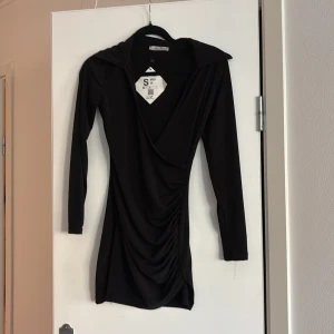 Svart omlottklänning från New Faveri - Snygg svart omlottklänning från New Faveri i storlek S. Klänningen har en elegant drapering och långa ärmar, perfekt för en stilren look.