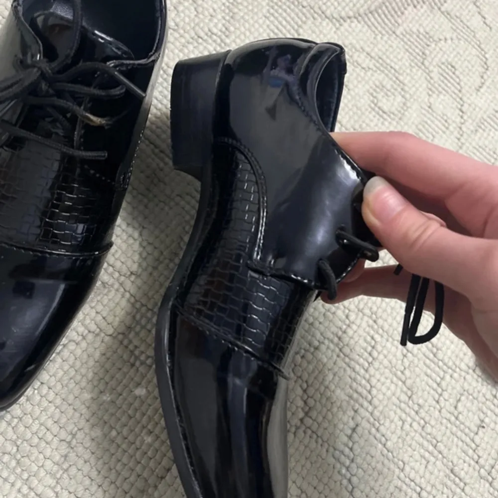 Säljer ett par svarta snörskor i lack med fyrkantig tå och låg klack. Skorna har ett stilrent mönster på ovandelen som ger en extra touch. Perfekta för dig som vill sticka ut med en klassisk men modern look. För barn.. Kengät.