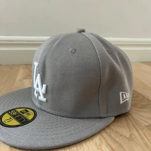 Grå LA-keps från New Era 59FIFTY - Snygg grå keps från New Era 59FIFTY med LA-logga broderad framtill i vitt. Klassisk modell med rak skärm och broderade ventilationshål. Perfekt för dig som gillar streetwear och sportig stil.