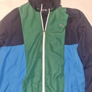 Lacoste jacka - Vindjacka från Lacoste i grönt och blått. Jackan har en dragkedja framtill och en huva med snörning. 